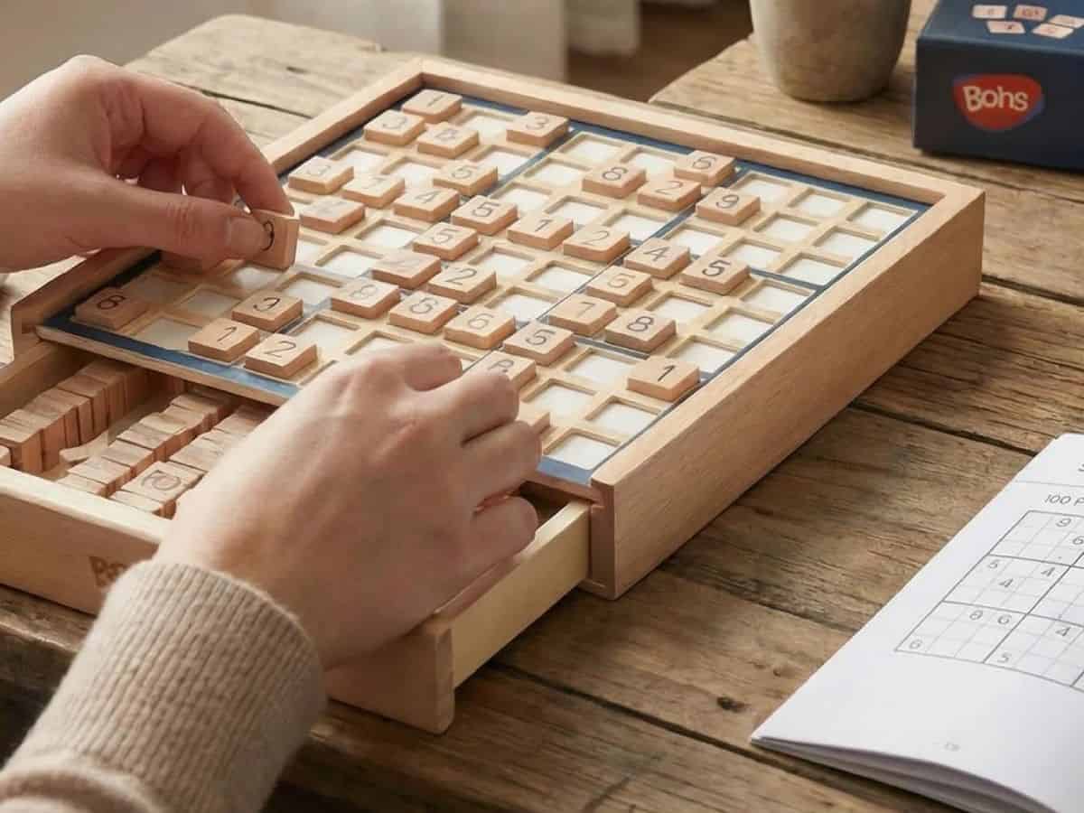 Due mani posizionano una tessera numerata sulla griglia del Sudoku in legno BOHS appoggiato su un tavolo, con il cassetto aperto e un libretto di puzzle accanto